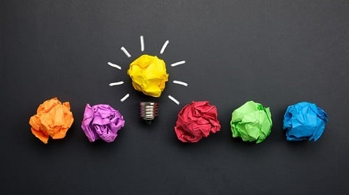 Un'immagine concettuale che rappresenta un'idea brillante che emerge da un processo creativo, visualizzata attraverso palline di carta colorata su uno sfondo grigio scuro.